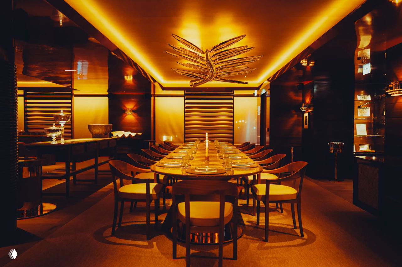 Интерьер приватной столовой ресторана La Palme d’Or: длинный стол, тёплое золотое освещение, элегантные стулья и крупная декоративная скульптура пальмовой ветви на потолке как центр композиции.