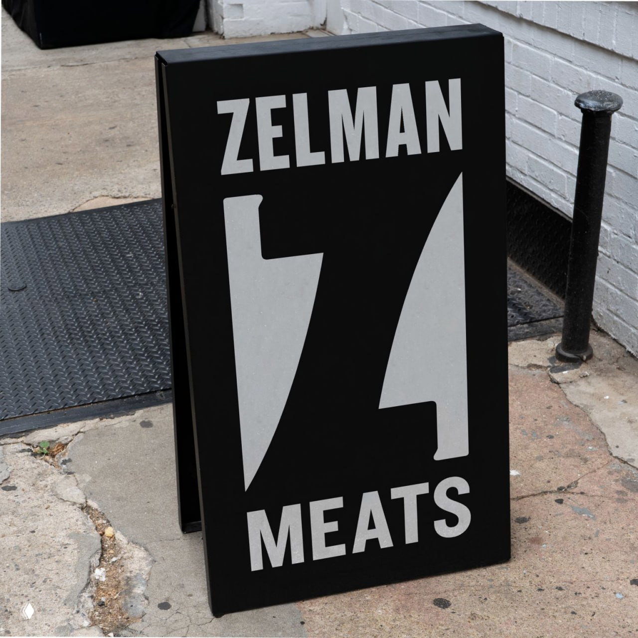 Черная уличная табличка с белым логотипом Zelman Meats, размещённая на бетонном тротуаре у входа — пример наружной навигации бренда.
