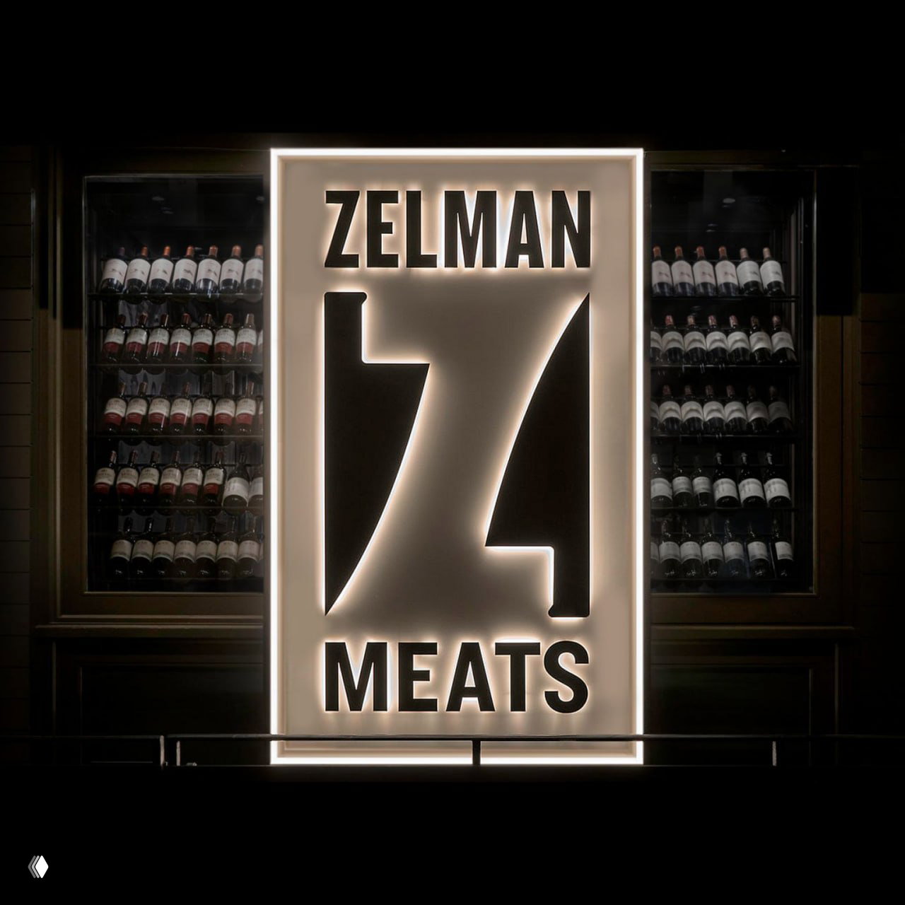 Подсвеченная вывеска Zelman Meats с крупным знаком: контрформа на световом коробе на фоне стены с винными бутылками, ночная съёмка.