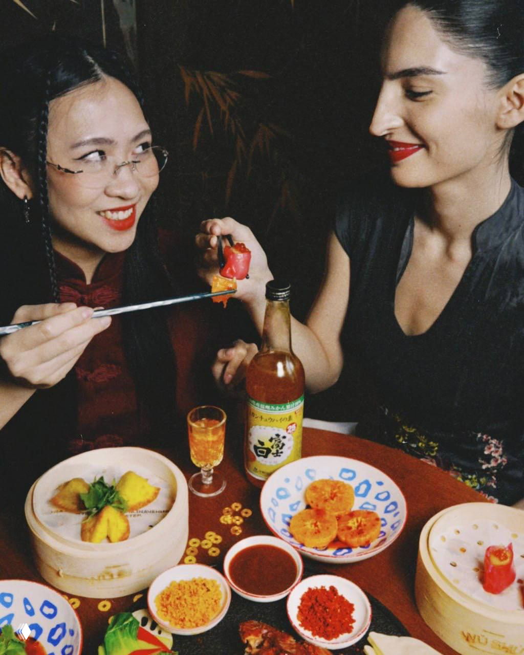 Две женщины за столом пробуют блюда из специального меню WU SHU: вонтоны, закуски, соусы и коктейли в тёплой праздничной обстановке.