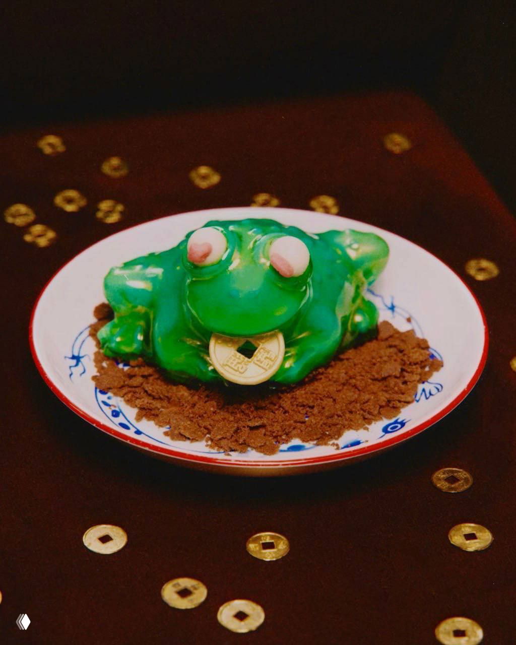 Десерт «гипно‑жаба» ярко‑зелёный с шоколадной монетой во рту, подан на тарелке с шоколадной крошкой и декоративными монетками вокруг.