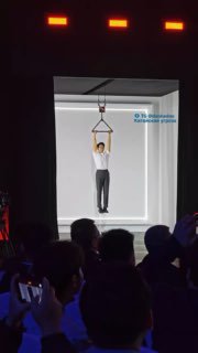 Фрагмент презентации Honor: демонстрация усиленного шарнира Magic V6 — человек держится за крюк в витрине, аудитория наблюдает на переднем плане