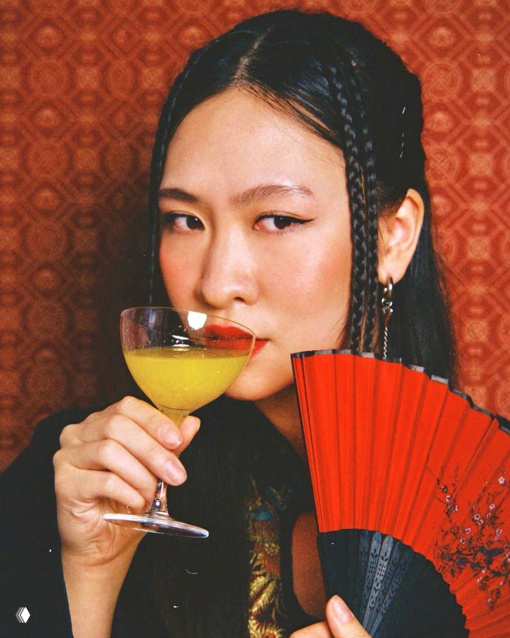 Женщина с жёлтым коктейлем и красным веером на фоне восточного узора, стилизованная фешн‑фотосессия в интерьере ресторана в духе праздника.