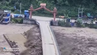 Разрушенный мост и затопленное русло реки: поток наносит грязь и камни, видны последствия оползня и перекрытия реки.