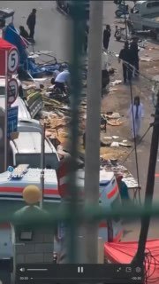 Кадр с места происшествия: скорые машины у палаток, разбросанные товары и люди рядом с пострадавшими, съёмка через ограждение или окно.