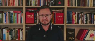 Видеокадр: мужчина среднего возраста в рубашке смотрит в камеру на фоне книжных полок; студийный фон, без идентификации.