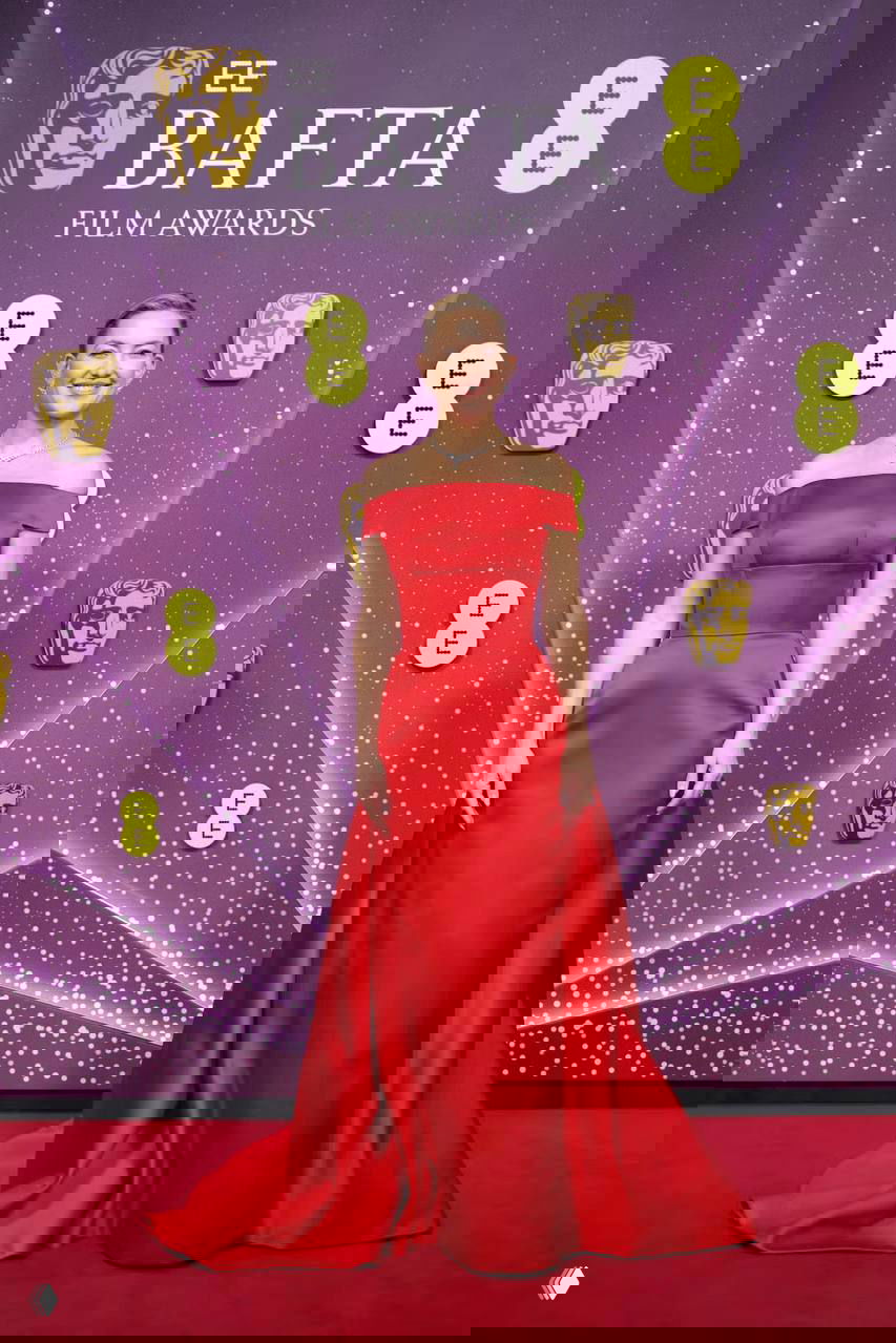 Женщина в ярком красном вечернем платье без бретелей позирует на красной дорожке BAFTA перед декорацией с символикой премии.