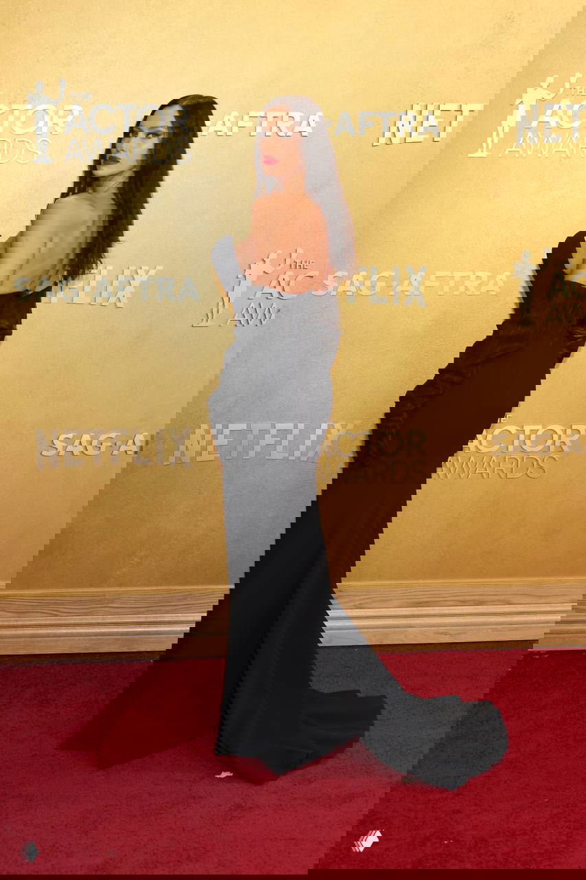 Задний план: чёрное платье со шлейфом и открытой спиной в позе на красной дорожке, золотая стена с надписями мероприятия на заднем фоне.