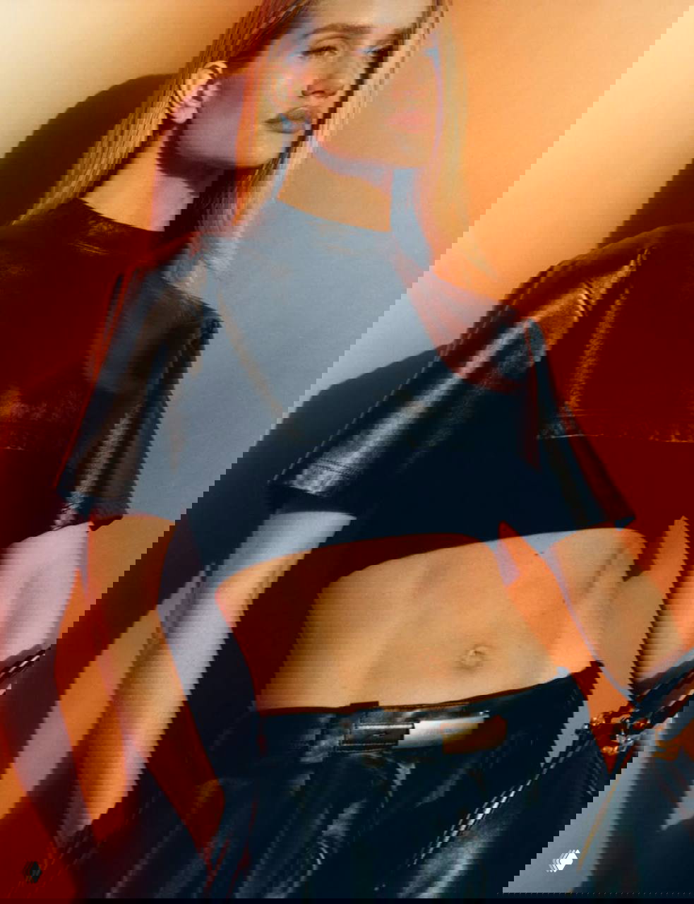 Фото в тёплой оранжевой палитре: модель в укороченном кожаном топе, виден пресс и профильная постановка тела под свет.