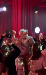 Гости окружают артистку, вручают большой букет цветов; кадр в розово-красной подсветке, видны смартфоны гостей, фиксирующие момент поздравления.