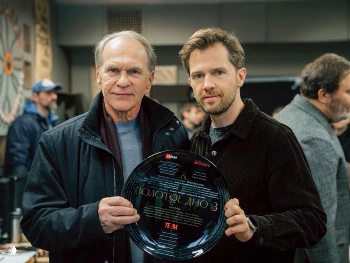Два мужчины на съёмочной площадке держат диск с надписью «ЗОЛОТОЕ ДНО 3», вокруг видна команда и съёмочный реквизит.
