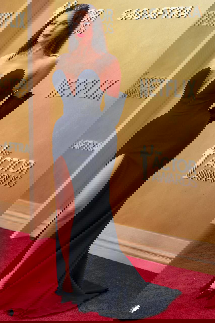 Портрет на красной дорожке: тёмные прямые волосы, яркий макияж и чёрное платье с характерным вырезом на фоне логотипов и ковра.