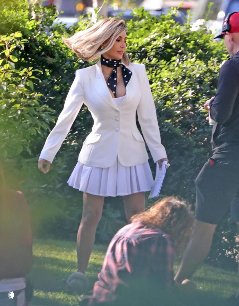 Женщина в белом костюме делает шаг по травяной площадке съёмочной площадки, в кадре видны ассистенты и оборудование.