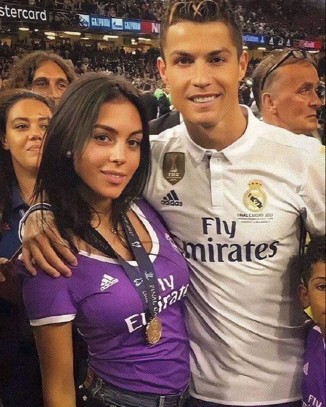 Криштиану Роналду в форме Real Madrid рядом с Джорджиной Родригес на трибуне стадиона, вокруг видна толпа болельщиков и праздничная атмосфера.