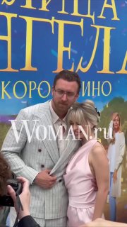 Крупный план: Данила Якушев обнимает супругу Кристину на фоне постера «Улица Ангела», видны фотографы и водяной знак Woman.ru
