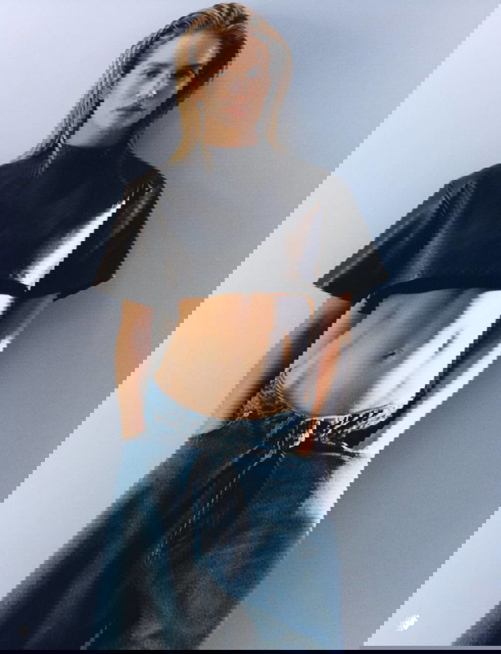 Вертикальный снимок модели в джинсовой юбке и кожаном топе, руки в карманах, естественная поза и дневной студийный свет.