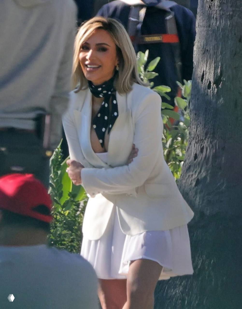 Кадр с открытой площадки: улыбающаяся женщина в белом пиджаке и юбке, вокруг люди и деревья, виден рабочий процесс съёмок.