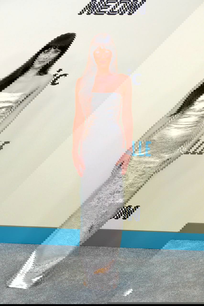 Женщина в длинном серебристом платье без бретелей, строгий силуэт и гладкая фактура ткани; образ на нейтральном светлом фоне красной дорожки.