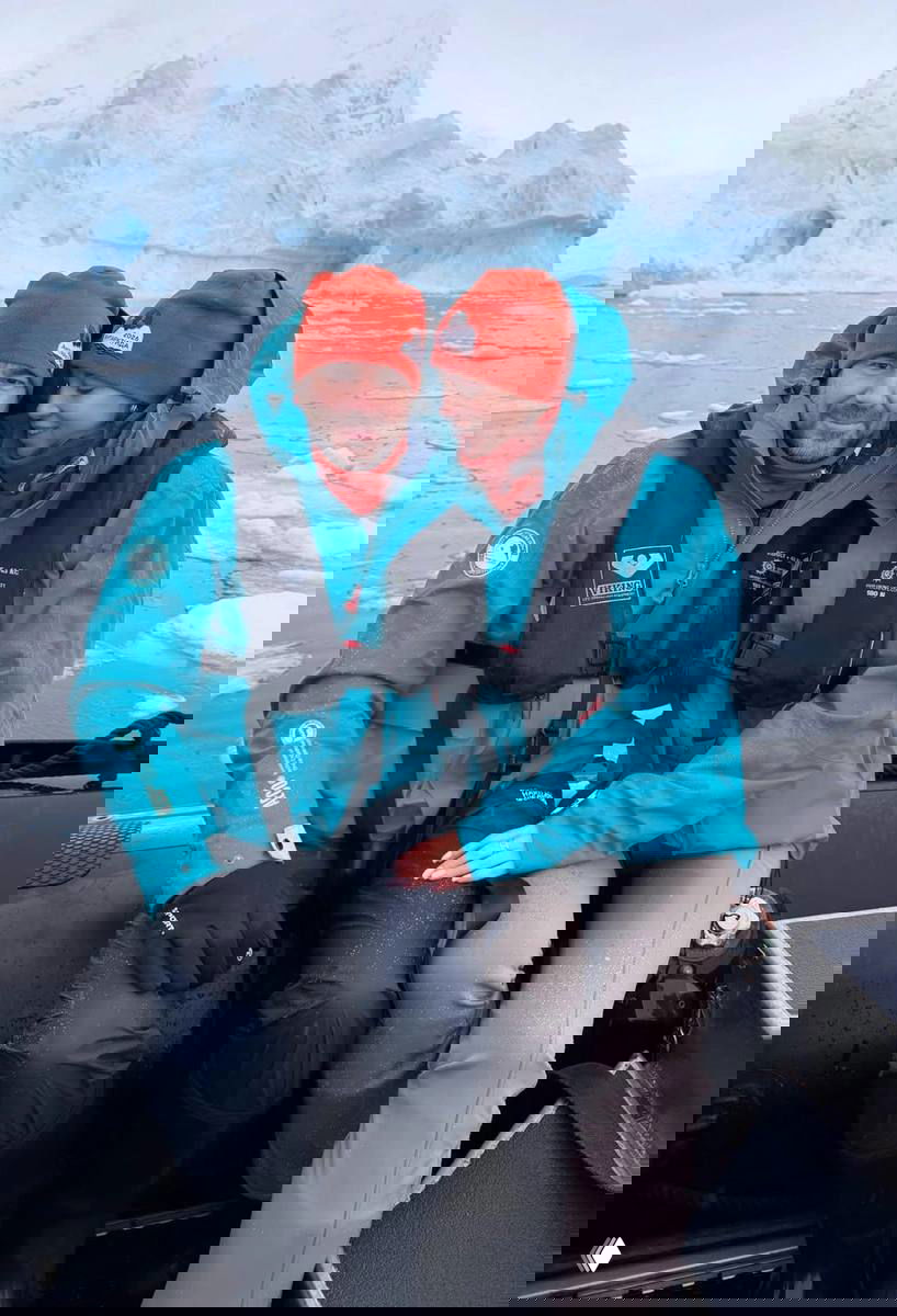 Пара в бирюзовых тёплых куртках и красных шапках сидит в резиновой лодке на фоне айсберга и ледяного пейзажа.