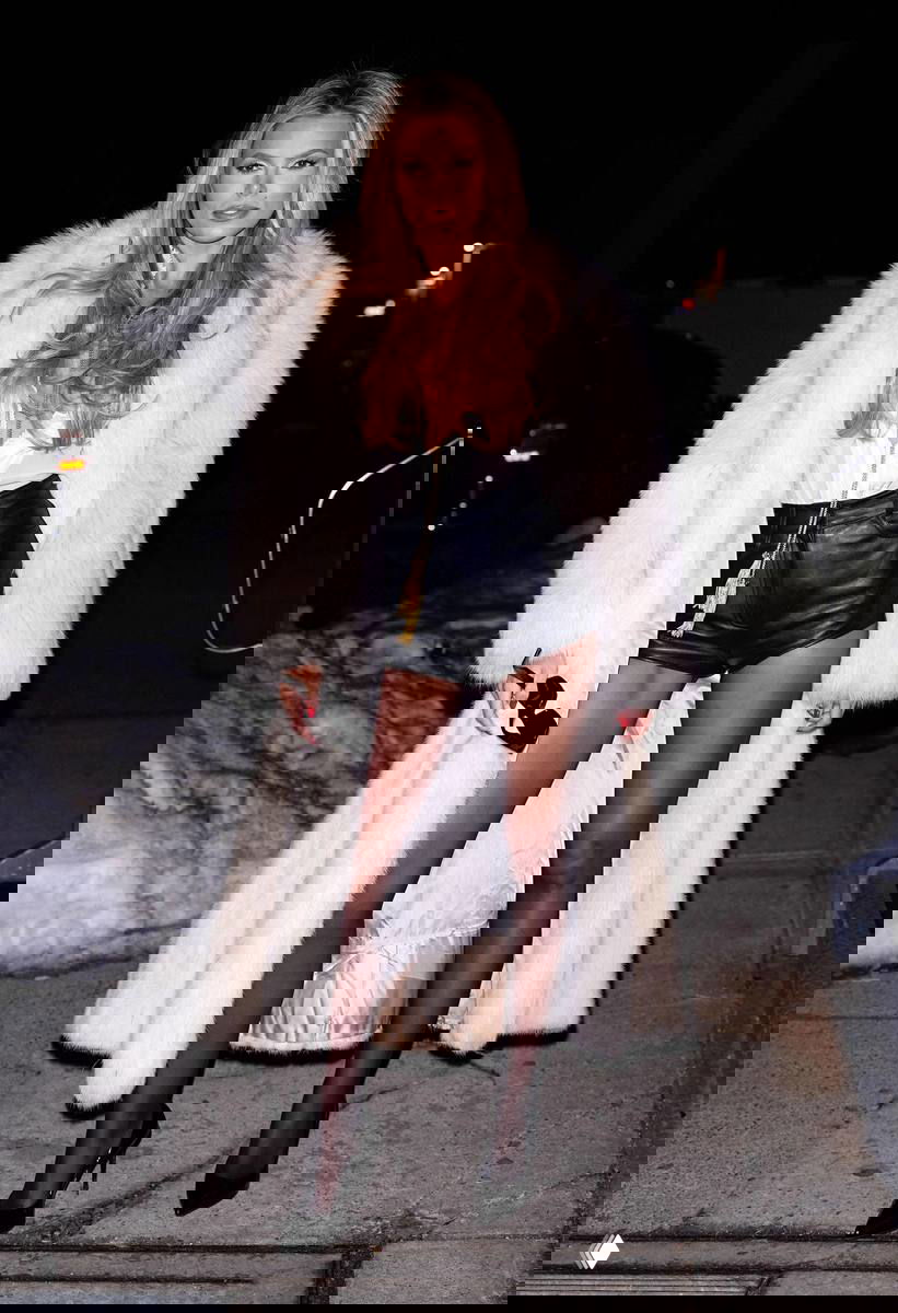 Женщина в длинной светлой шубе и кожаных шортах идёт по ночной улице, на тротуаре виден снег и припаркованные автомобили.