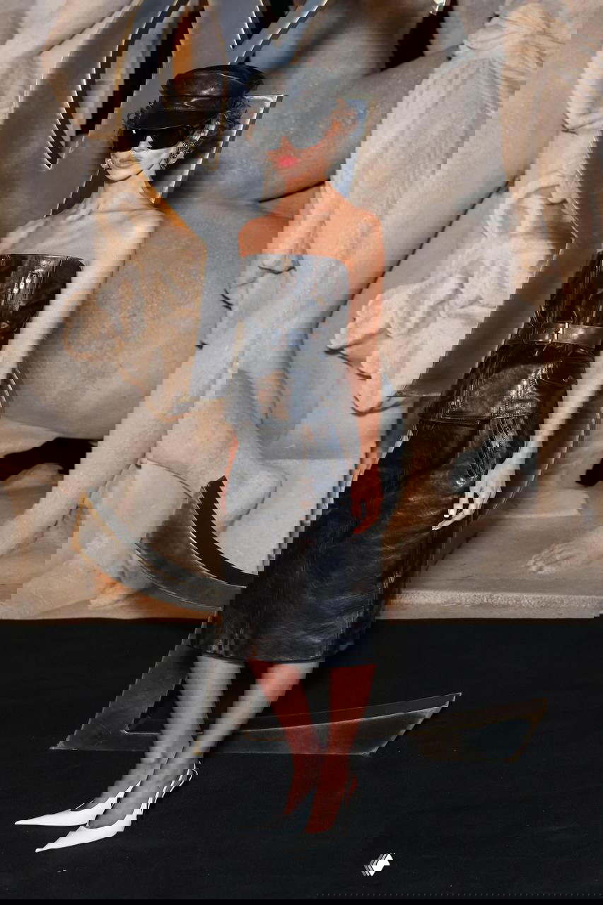 Модель в чёрном кожаном корсетном топе и юбке с чёрной кепкой и крупными очками на фоне металлического логотипа Saint Laurent во время шоу.