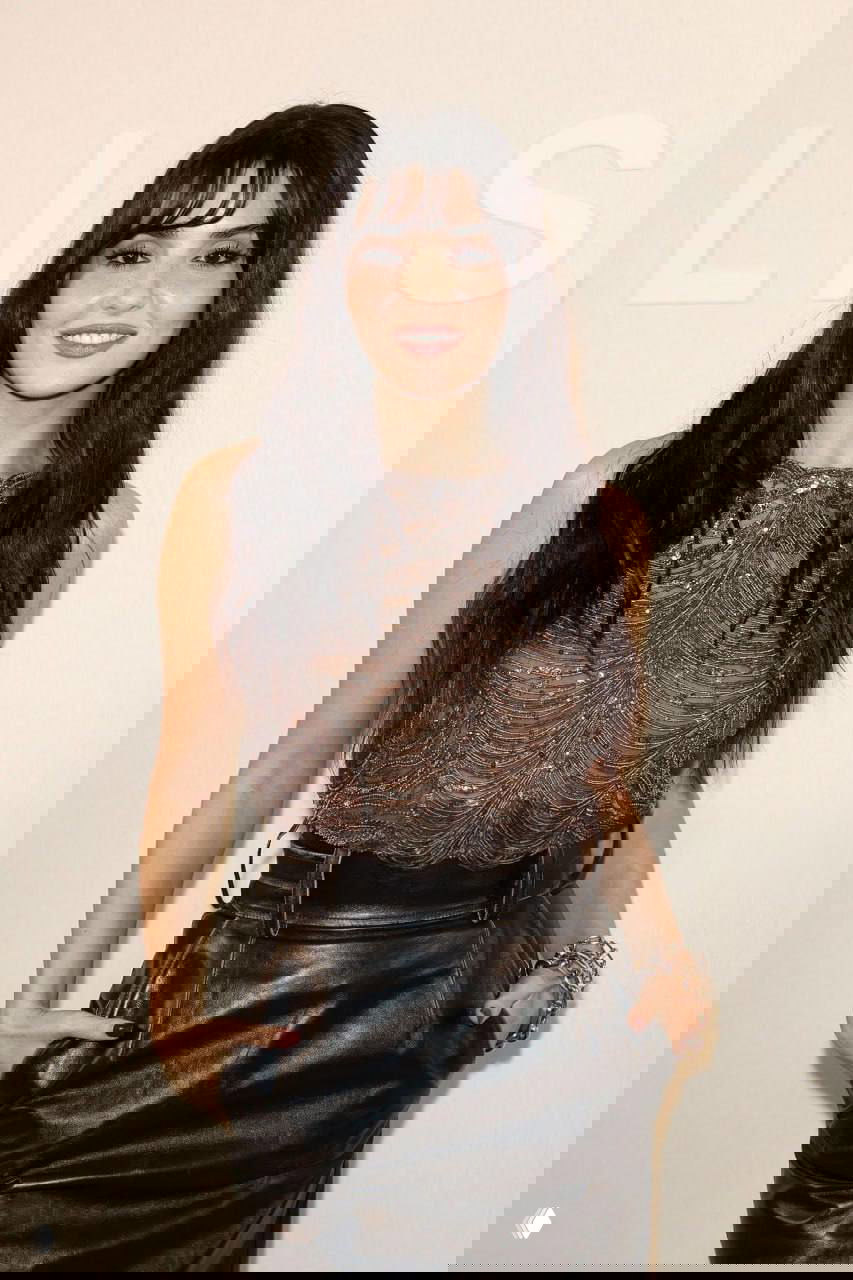 Ханде Эрчел позирует у стены бренда Elie Saab в Москве: вечерний топ с драпировкой, кожаная юбка и украшения на светлом фоне под логотипом бренда.