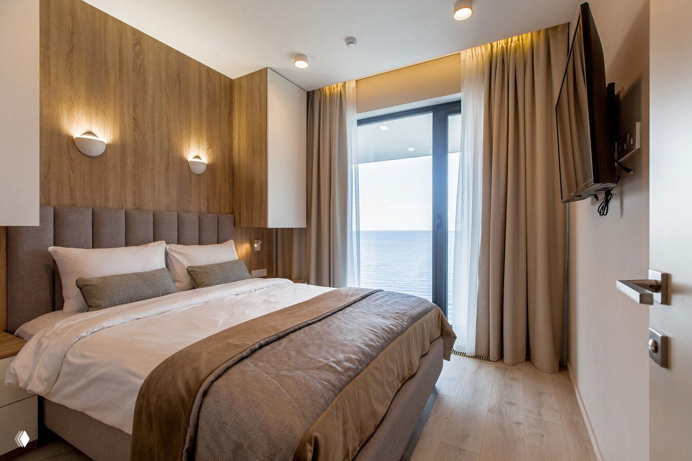 Спальня с большой кроватью и панорамным окном, через которое открывается вид на море и балкон, светлая текстильная отделка.