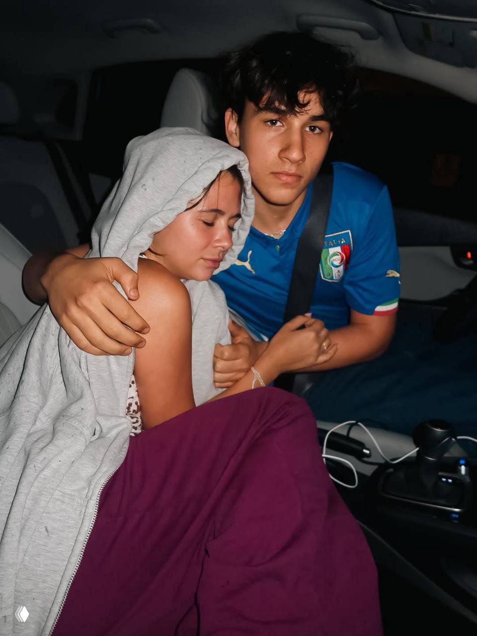 Ночной снимок в салоне автомобиля: подросток в синей футболке обнимает девушку в худи, видно ремень безопасности и часть интерьера машины.