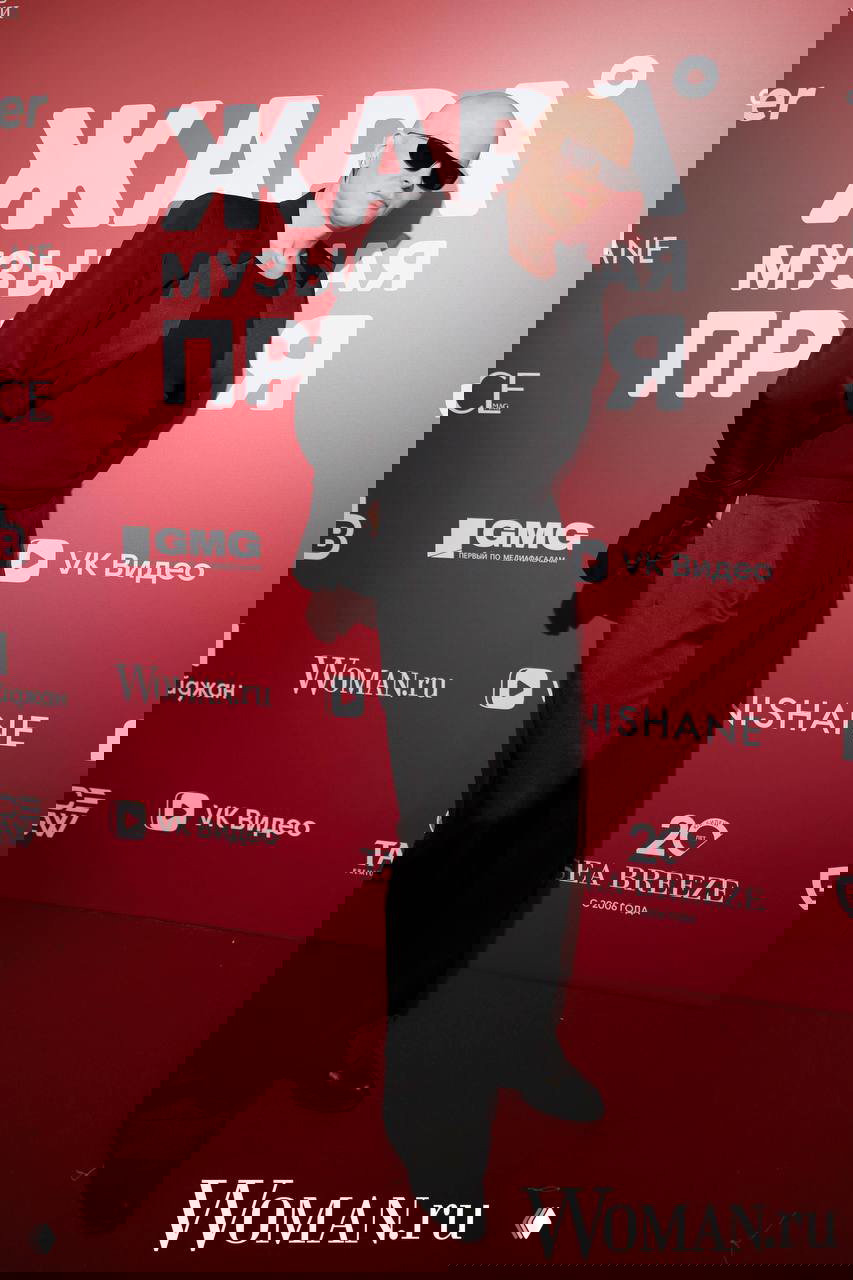 Высокий мужчина в тёмном костюме и солнцезащитных очках позирует на красной дорожке перед фотостеной с крупной надписью названия премии.