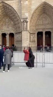 Сцена возле соборного входа в Париже: люди и фотографы у ограждений, на переднем плане женщина с сумкой и городской пейзаж.