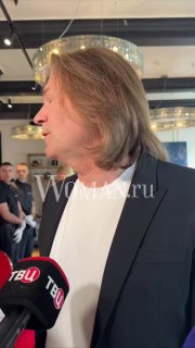 Профиль мужчины среднего возраста в пиджаке даёт комментарий у микрофонов журналистов в общественном помещении.