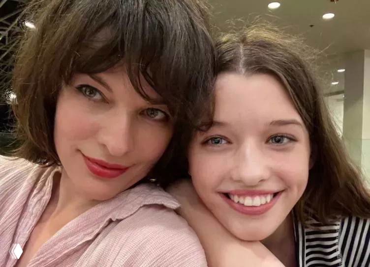 Крупный план женщины и молодой девушки, улыбаются в селфи-стиле; естественное выражение лица, домашняя обстановка и мягкое освещение.