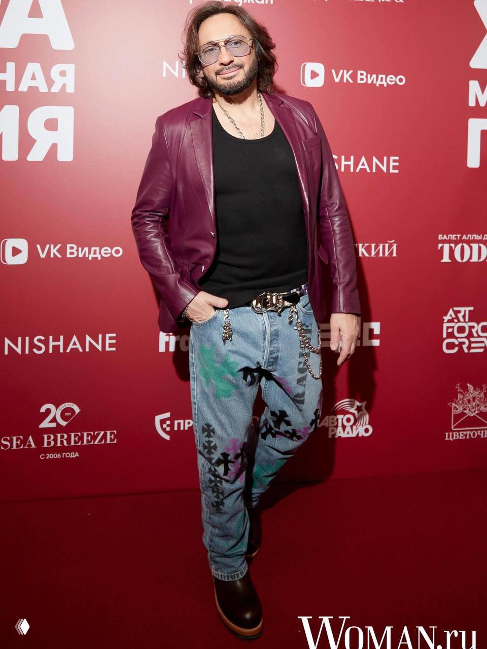 Мужчина в бордовой кожаной куртке и расписанных джинсах позирует на красной дорожке рядом с логотипом Woman.ru на фоне пресс-волла.