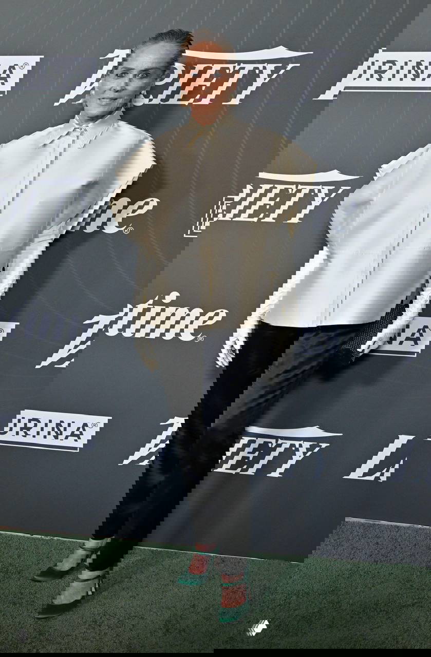 Женщина в светлой короткой блузе с длинными перчатками и тёмных атласных брюках стоит на фоне логотипов Variety, образ в стиле минимализма.