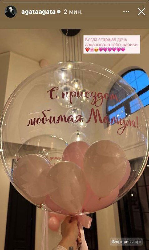 Прозрачный шар с розовыми воздушными шарами внутри и надписью «С приездом, любимая мамуля», держат рукой на фоне интерьера — подарок от старшей дочери.