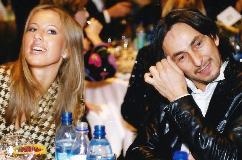 Двое гостей за столом на торжественном ужине: мужчина в чёрной кожаной куртке и женщина слева, на переднем плане бутылки с водой и праздничная обстановка.