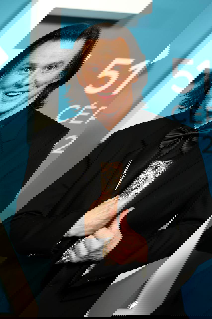 Крупный портрет мужчины в чёрном смокинге на фоне синей фотостены церемонии: улыбка, бант и держимая статуэтка в руках вечером в Париже