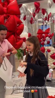 Девочка получает подарок и улыбается на фоне стены из шаров‑сердец и брендовых коробок, вокруг видно праздничные аксессуары и игрушки.