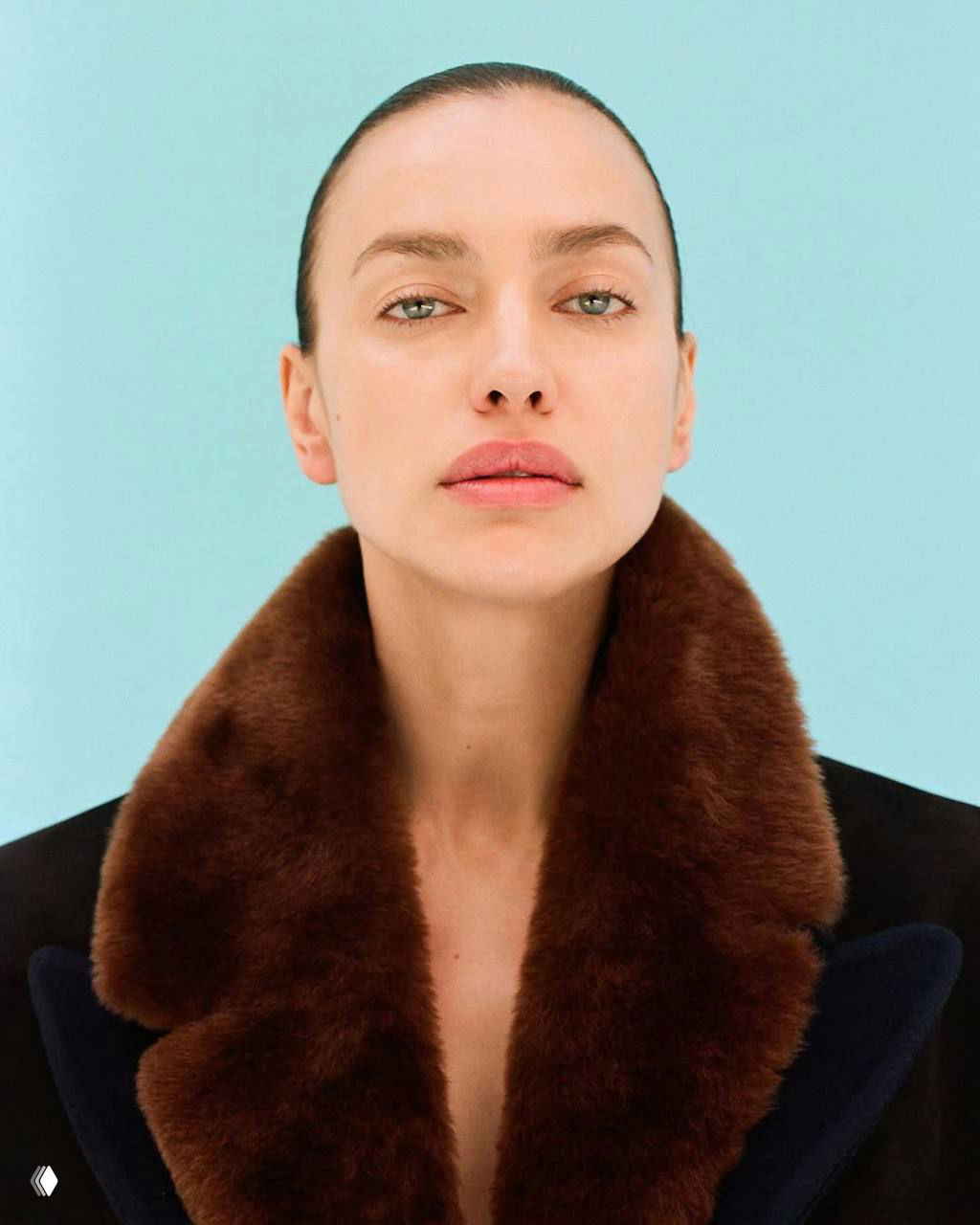 Крупный портрет Ирины Шейк на светло-голубом фоне в меховом воротнике — натуральный макияж и спокойный взгляд в камеру.