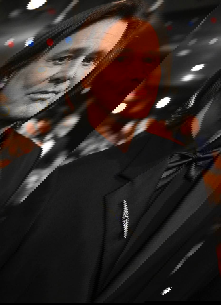 Полуфронтальный портрет мужчины в смокинге на церемонии: спокойное выражение лица, освещение камер и расфокусированная публика позади