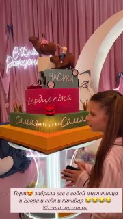 Многоярусный праздничный торт со свечами и декоративными табличками на столе; девочка наклоняется над тортом в момент поздравления