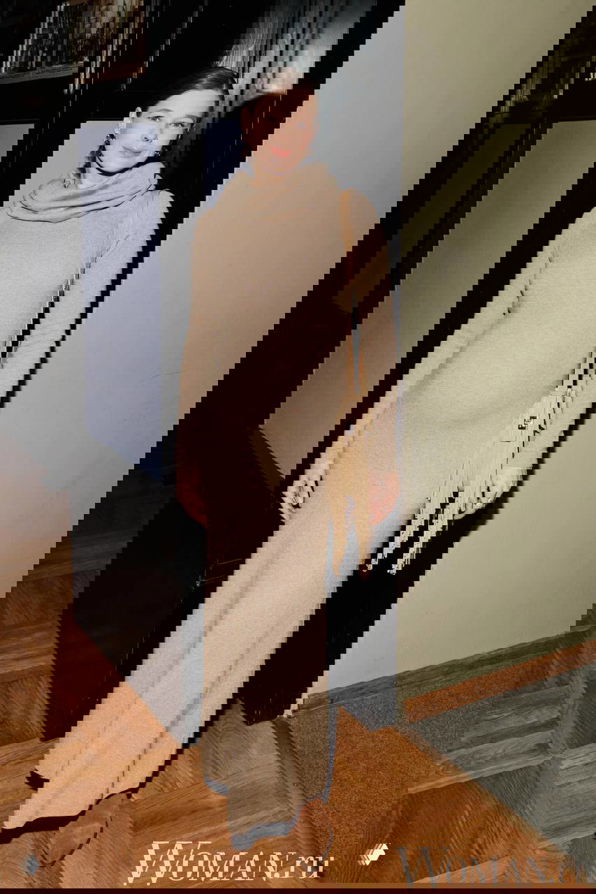 Женщина в длинном бежевом трикотажном платье и замшевых ботинках, с плечевой сумкой, позирует в коридоре театра — снимок с церемонии.