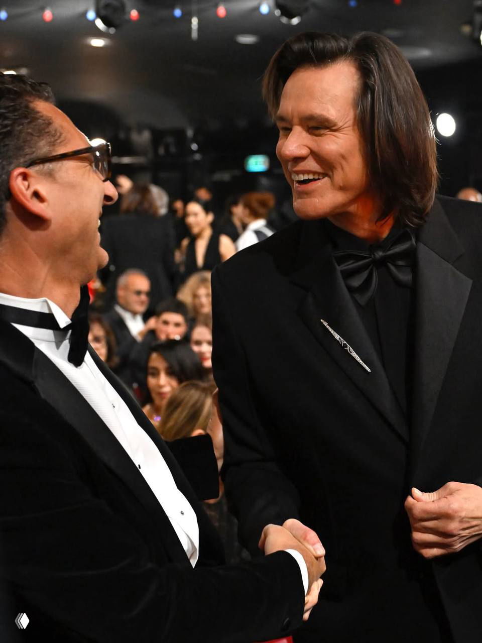 Мужчина в чёрном смокинге пожимает руку коллеге в смокинге во время церемонии: живое общение в зале с зрителями на заднем плане