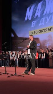 Человек на сцене с микрофоном перед большим экраном, на экране видна рекламная графика с текстом и датой релиза фильма.