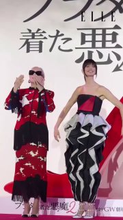 Две женщины на промо-туре в Токио в эффектных дизайнерских образах: одна в красно-чёрном платье с бахромой и очками, вторая — в чёрно‑белом волнообразном платье.