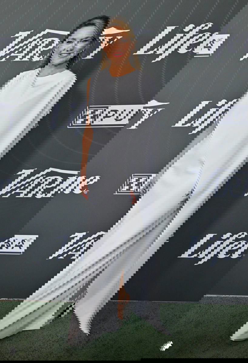 Женщина в длинном белом платье с драпировкой и высоким разрезом позирует перед пресс-воллом Variety, элегантный вечерний лук.