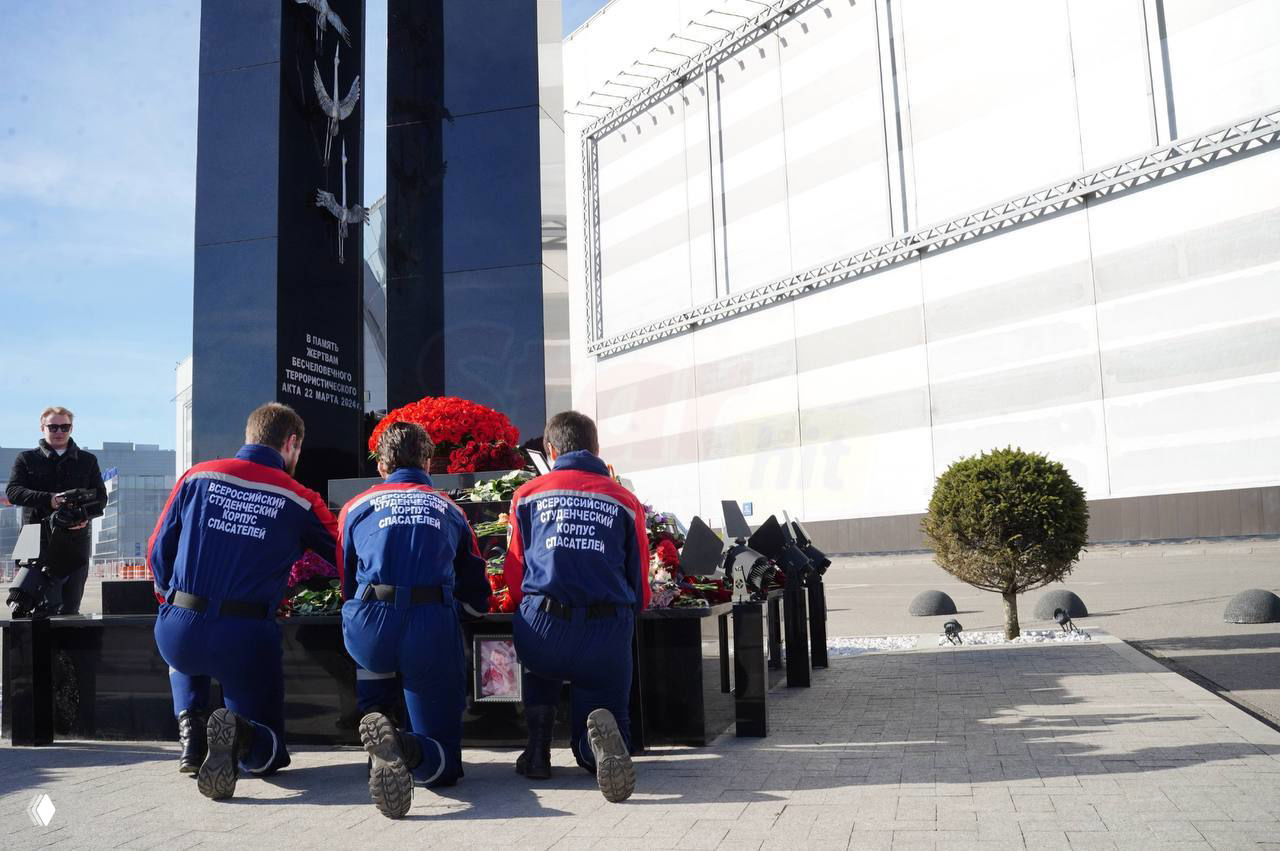 Три человека в форме спасателей преклонили колени перед монументом и венками у мемориала, скорбная церемония возложения цветов