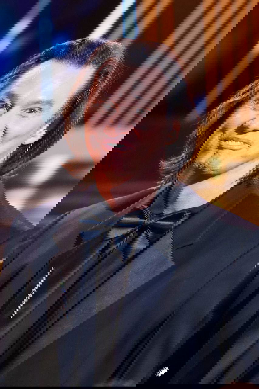 Ещё один портрет с красной ковровой дорожки: мужчина в смокинге с лёгкой улыбкой, крупный план под сценическими прожекторами и залом позади