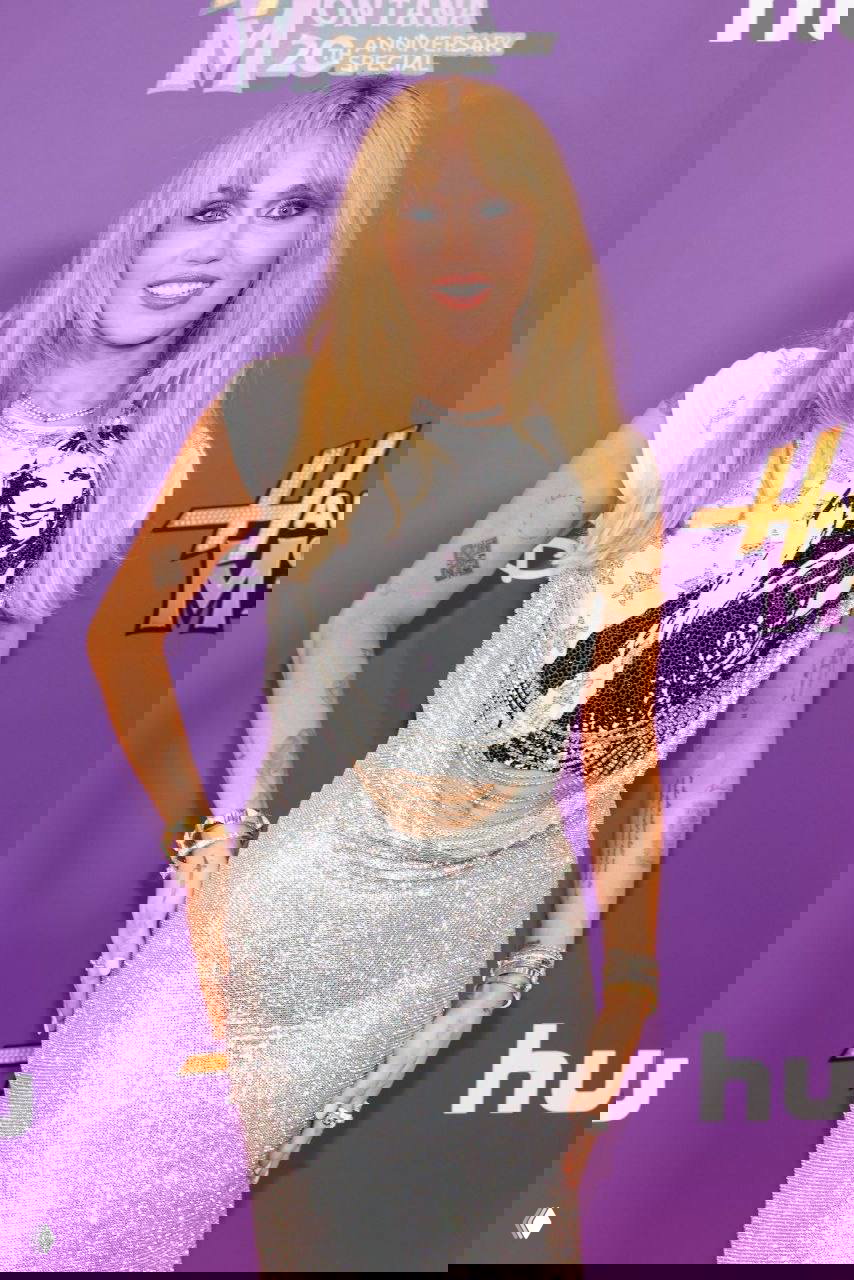 Майли Сайрус в полный рост на фиолетовой ковровой дорожке: серебристая юбка, футболка с принтом и фон с логотипом «Hannah Montana».