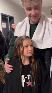 Девочка стоит рядом с мужчиной за кулисами в коридоре концертного пространства: тёплый кадр перед концертом, без указания личности взрослого.